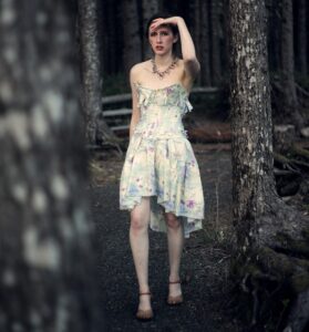 a beautiful young woman in a white sundress stands between two trees with a hand above her eyes as though she is searching for something.
